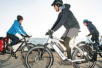 Drei Personen fahren auf E-Bikes an einem Seeufer.