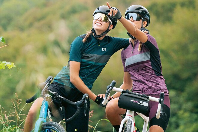 Neuheiten 2023. Zwei Frauen mit Fahrradtricots und Helmen stehen vor einem grünen Hintergrund.