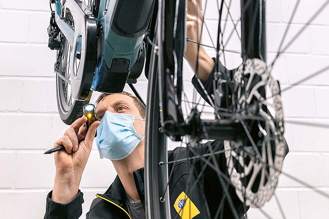 Ein Mann mit Mundnasen-Schutz leuchtet mit einer Taschenlampe auf den Motor eines E-Bikes