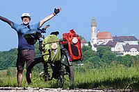 Ein Mann steht neben einem Tourenrad mit Packtaschen und hebt die Arme in die Luft.