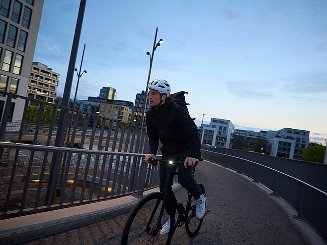 Mann mit weissem Uvex Velohelm fährt in der Dämmerung einer Brück entlang