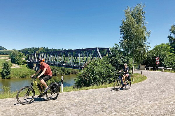 Locker durch die Schweiz radeln: Vier Routen zwischen Bodensee und Genfersee. Ein Mann und eine Frau auf E-Bikes, im Hintergrund ein Fluss.