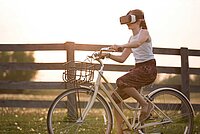 Mädchen mit VR-Brille fährt auf dem Fahrrad an einem Holzzaun entlang.