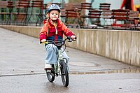 Ein Kind mit roter Jacke und Fahrradhelm fährt auf einem Kindervelo.