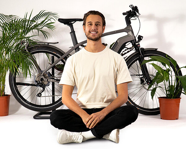 Ein Mann mit weissem T-Shirt sitzt im Schneidersitz vor einem Fahrrad.