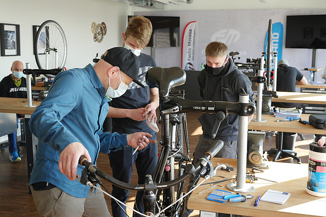Drei Männer mit Nasenmundschutz führen Servicearbeiten an einem Fahrrad aus.
