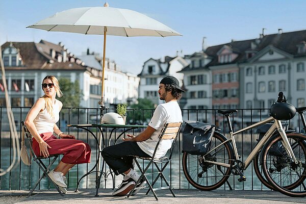Eine Frau und ein Mann sitzen an einem Bistrotisch am Ufer eines Flusses.