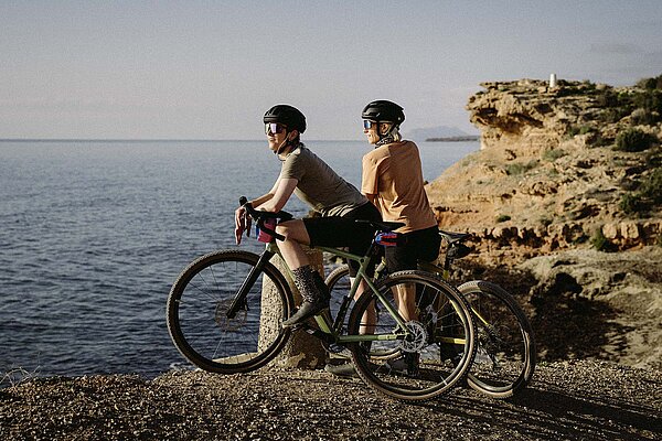 Zwei Rennradfahrer stehen an einer Küste und blicken aufs Meer.