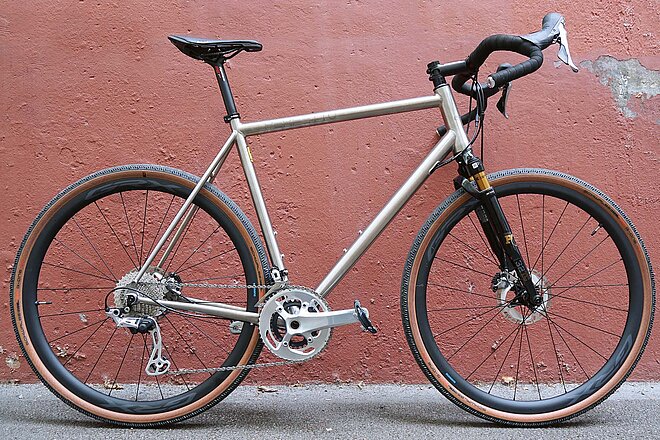 Citycycles Perfetto Gravelbike. Gravelbike aus Titan vor roter Mauer. 