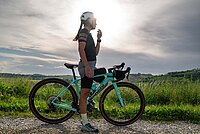 Bianchi Arcadex Gravelbikes. Eine Frau steht neben ihrem Gravelbike. Dahinter eine grüne Wiese.