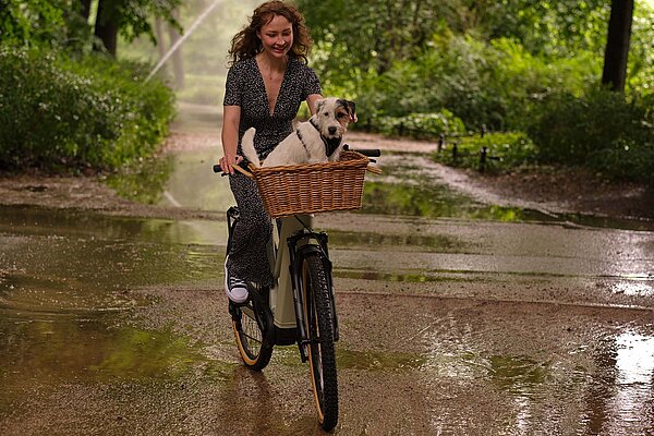 Tipps zum Kauf eines Occasions E-Bikes. Eine Frau mit Jumpsuit fährt auf einem E-Bike. Vorne ein Hund in einem Korb.