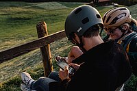Zwei Personen mit Fahrradhelm sitzen auf einer Bank und schauen auf ein Smartphone.