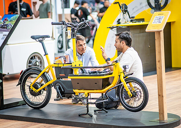 Zwei Männer knien neben einem gelben Lastenrad am Boden. 