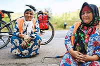 Zwei Frauen in volkstümlicher Kleidung kauern am Boden, hinter ihnen steht ein Elektrovelo